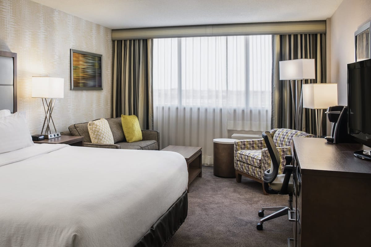 A hotel room with a large bed, a desk, a chair, a small table, and a TV.