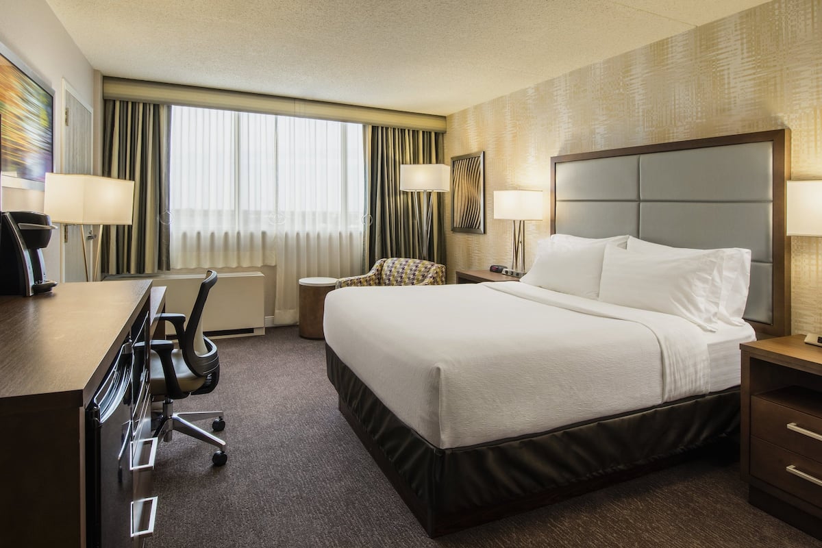 A hotel room with a bed, desk, chair, TV, lamps, and a window with curtains.