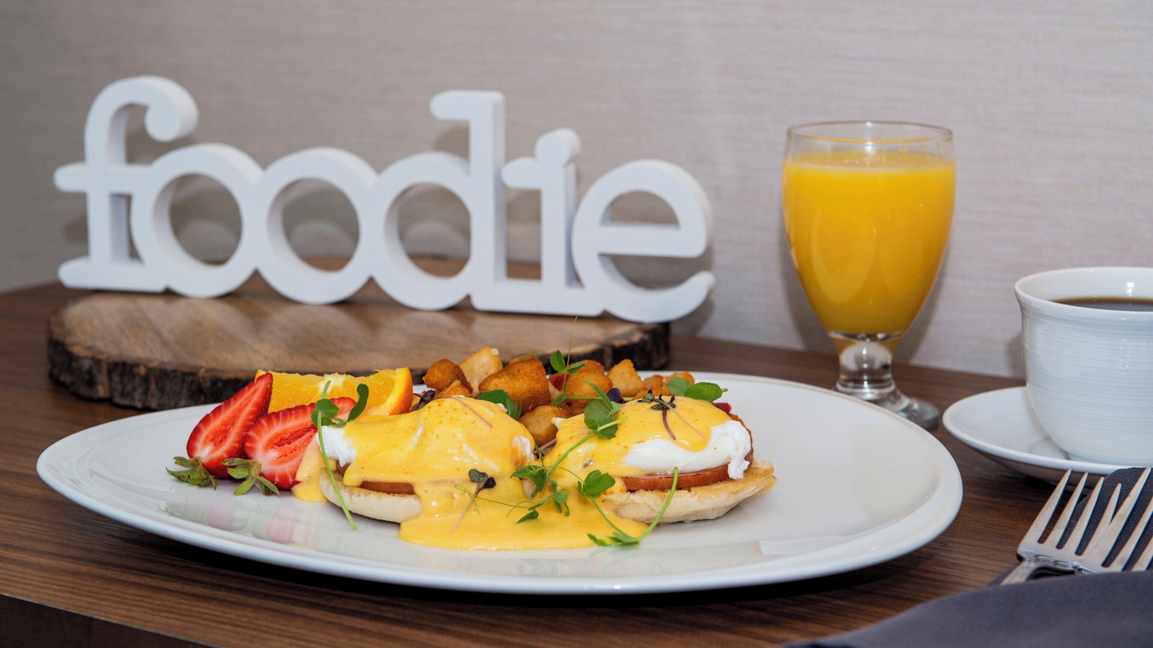 Een ontbijt bord met Eggs Benedict, toast en fruit, een glas sinaasappelsap en een kop koffie op een houten tafel.