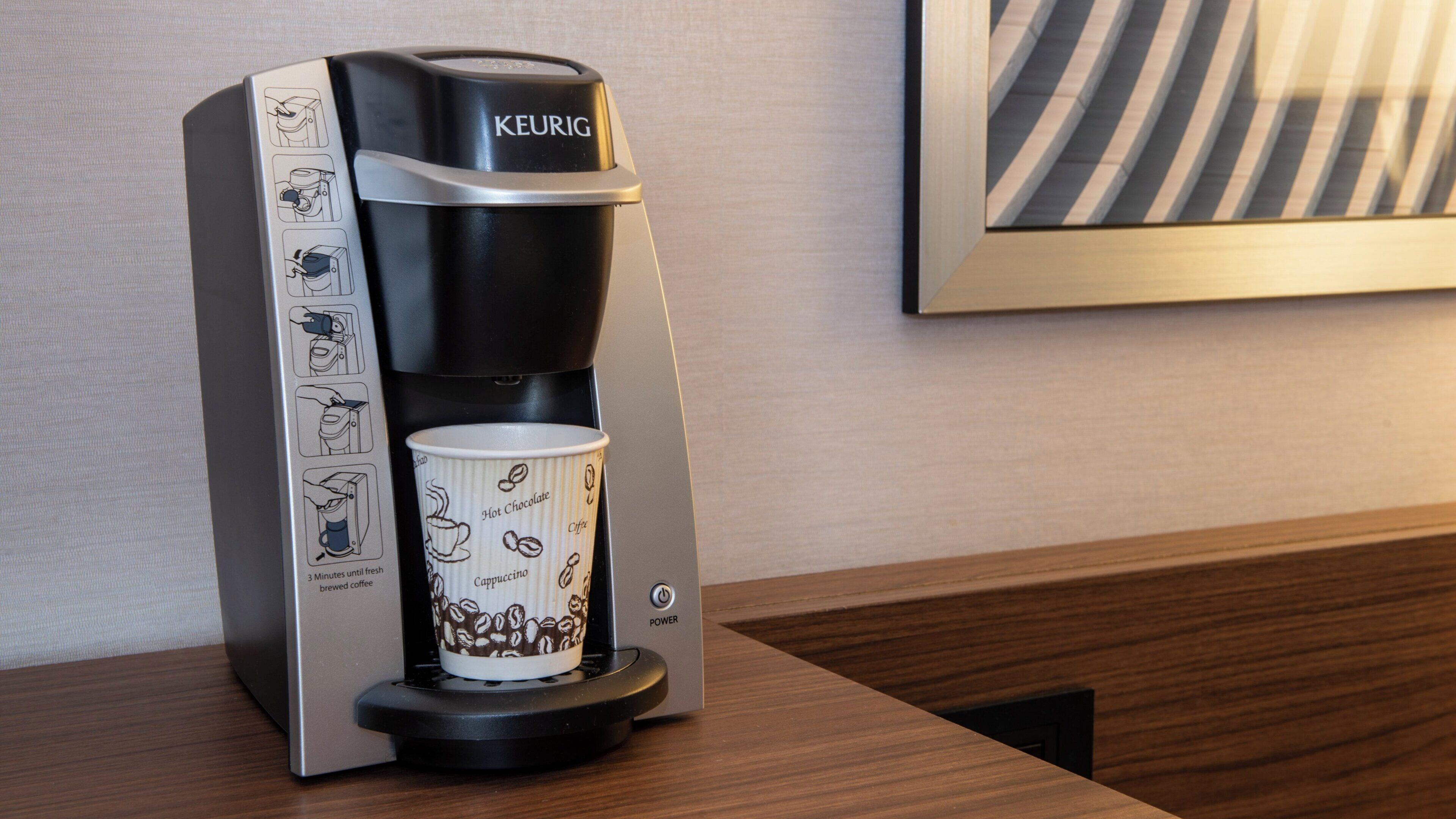 Una cafetera Keurig con una taza de café sobre la encimera, instrucciones a un lado y un cuadro en la pared.