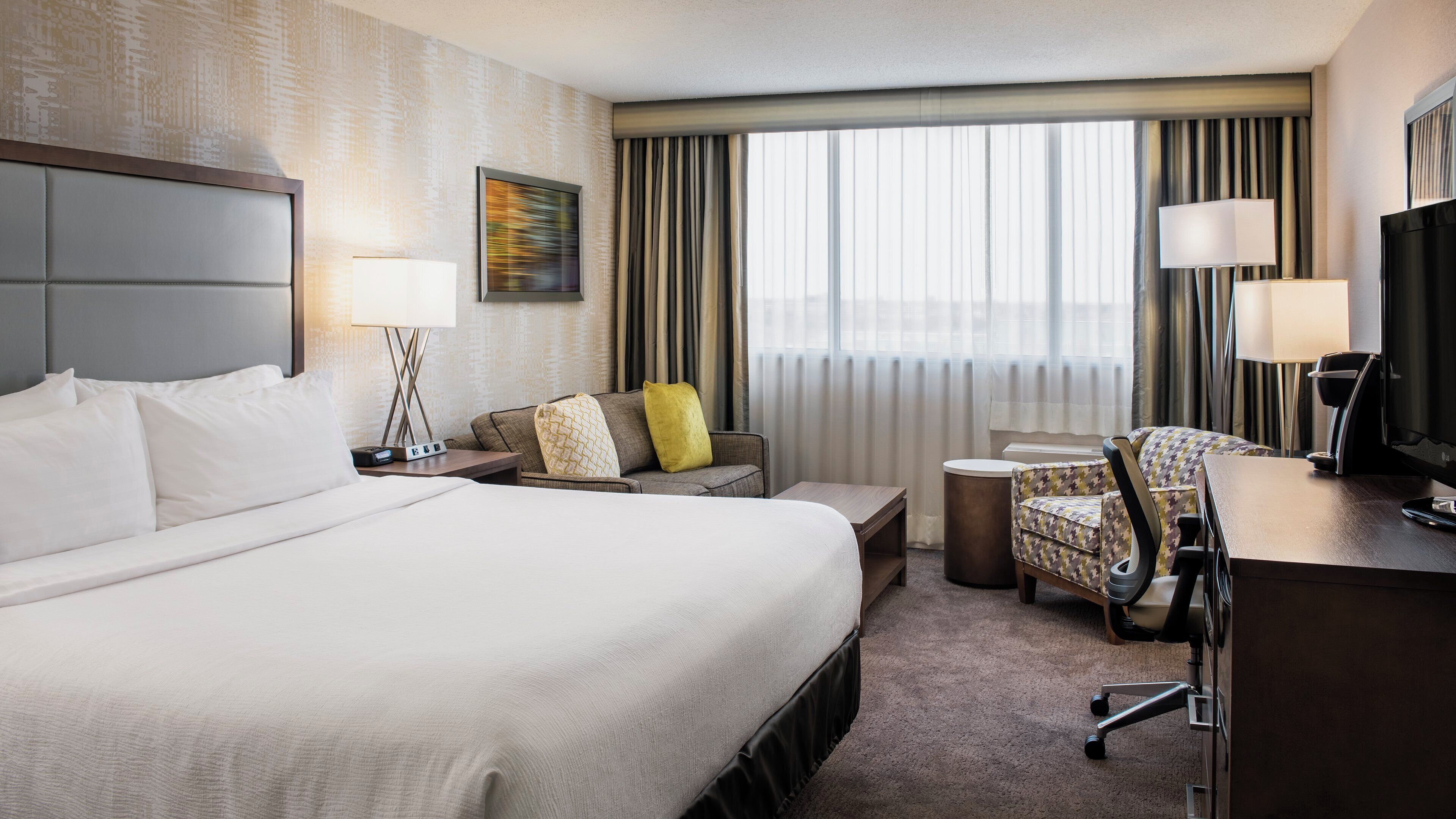 Habitación de hotel con una cama grande, un escritorio con silla, televisión y ventana con cortinas.