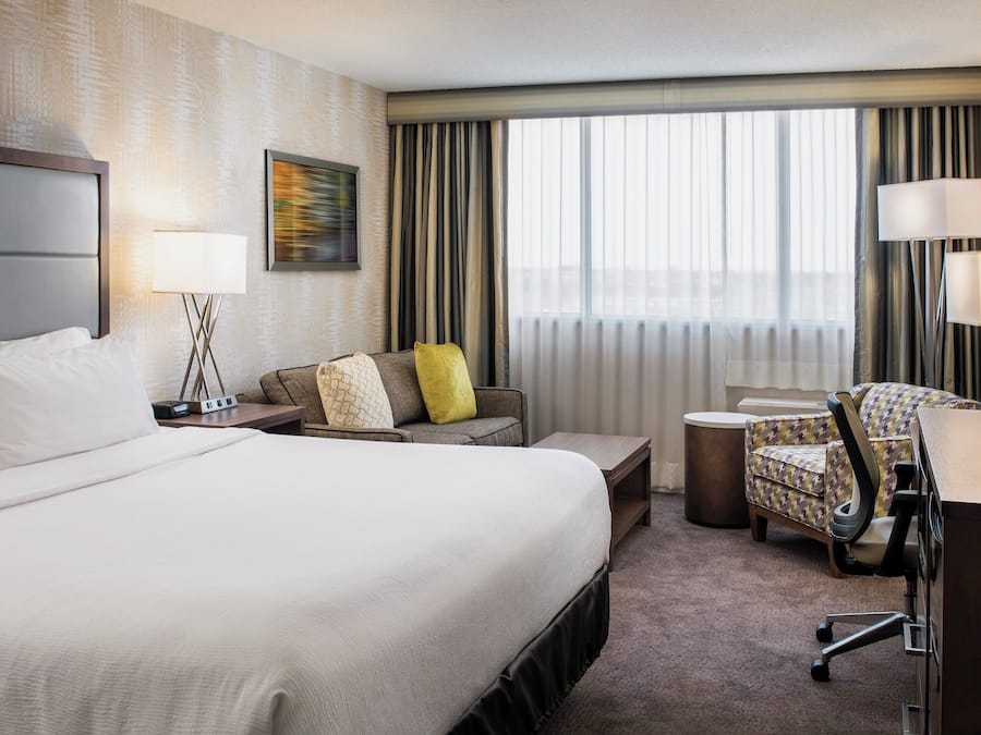 Habitación de hotel con una cama grande, un escritorio con silla, televisión y ventana con cortinas.