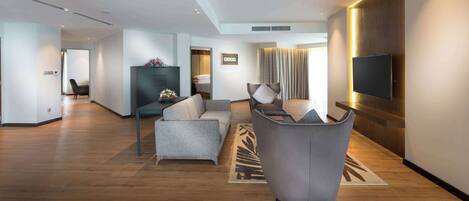 Habitación de hotel moderna con sofá gris, sillón gris, televisor de pantalla plana en la pared y piso de madera.