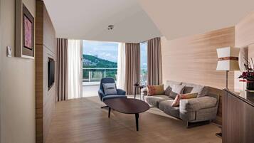 A modern hotel room with a wooden floor, a sofa, a chair, a TV, and a balcony with a view of a green hill and buildings.