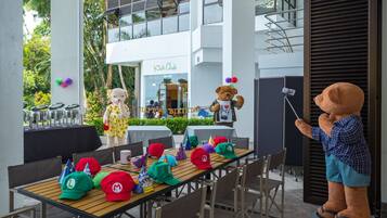 An outdoor dining area with a table set for a birthday party, featuring a teddy bear mascot and a children's club building in the background.
