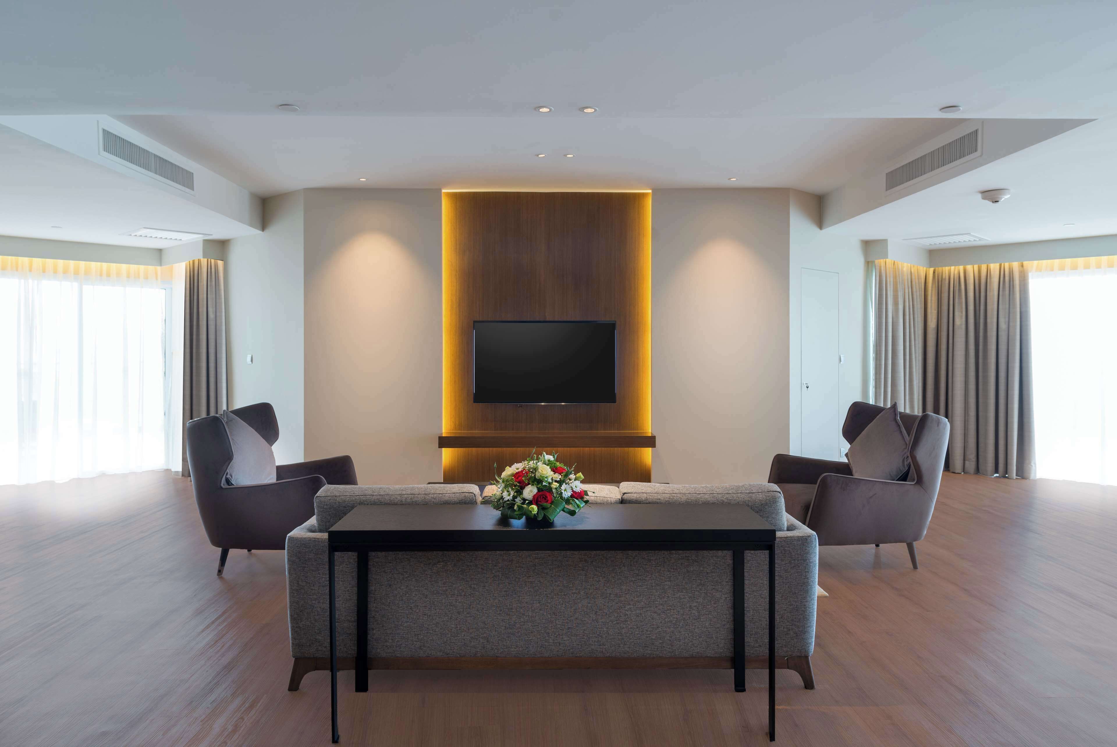 A modern living room with a flat-screen TV mounted on a wooden panel, two armchairs, a coffee table with a floral centerpiece, and large windows with curtains.