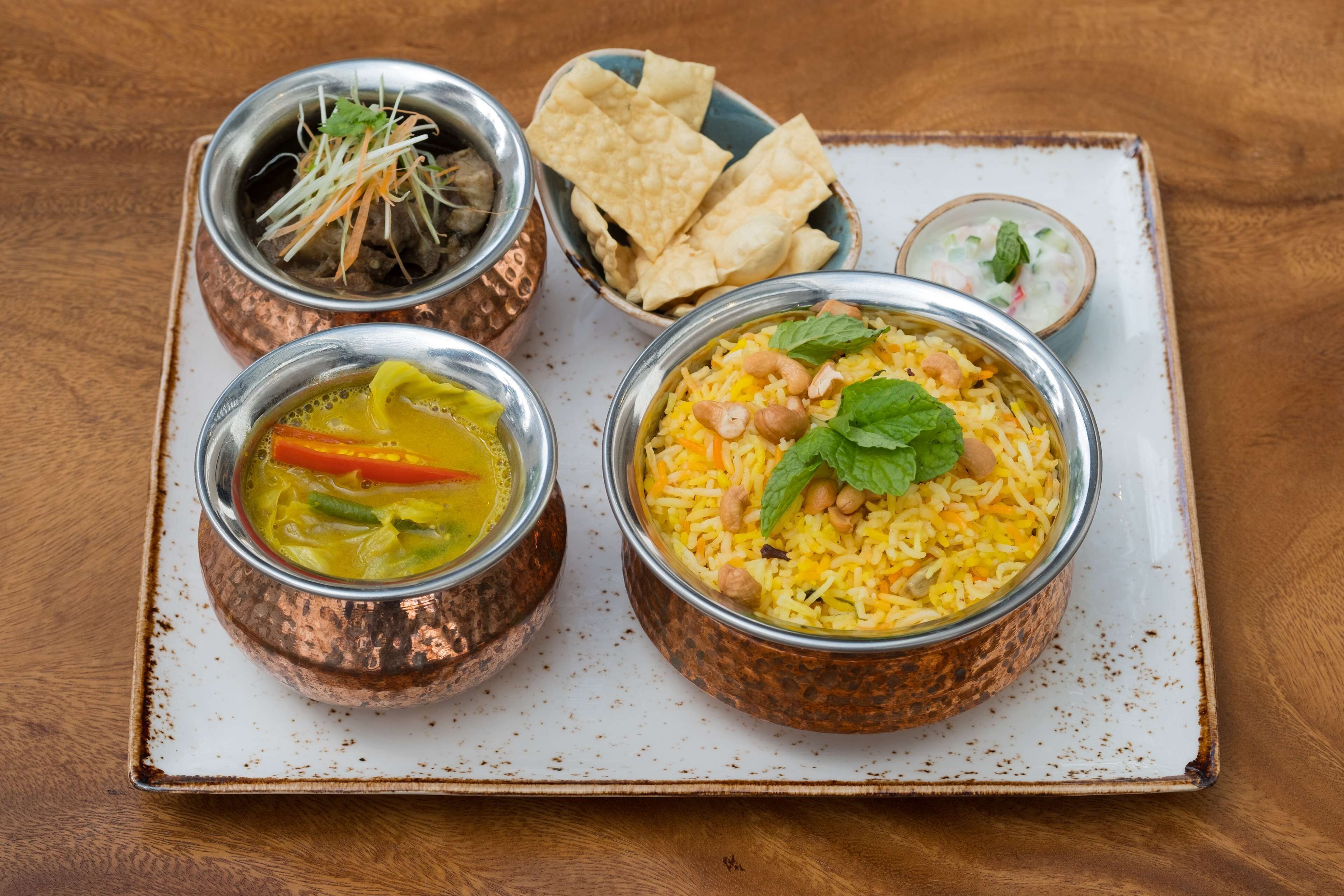 Una bandeja con dos tazones de arroz, uno con arroz amarillo adornado con nueces y menta, y otro con arroz amarillo y verduras. También hay dos recipientes pequeños, uno con un plato de verdura verde y otro con sopa amarilla.