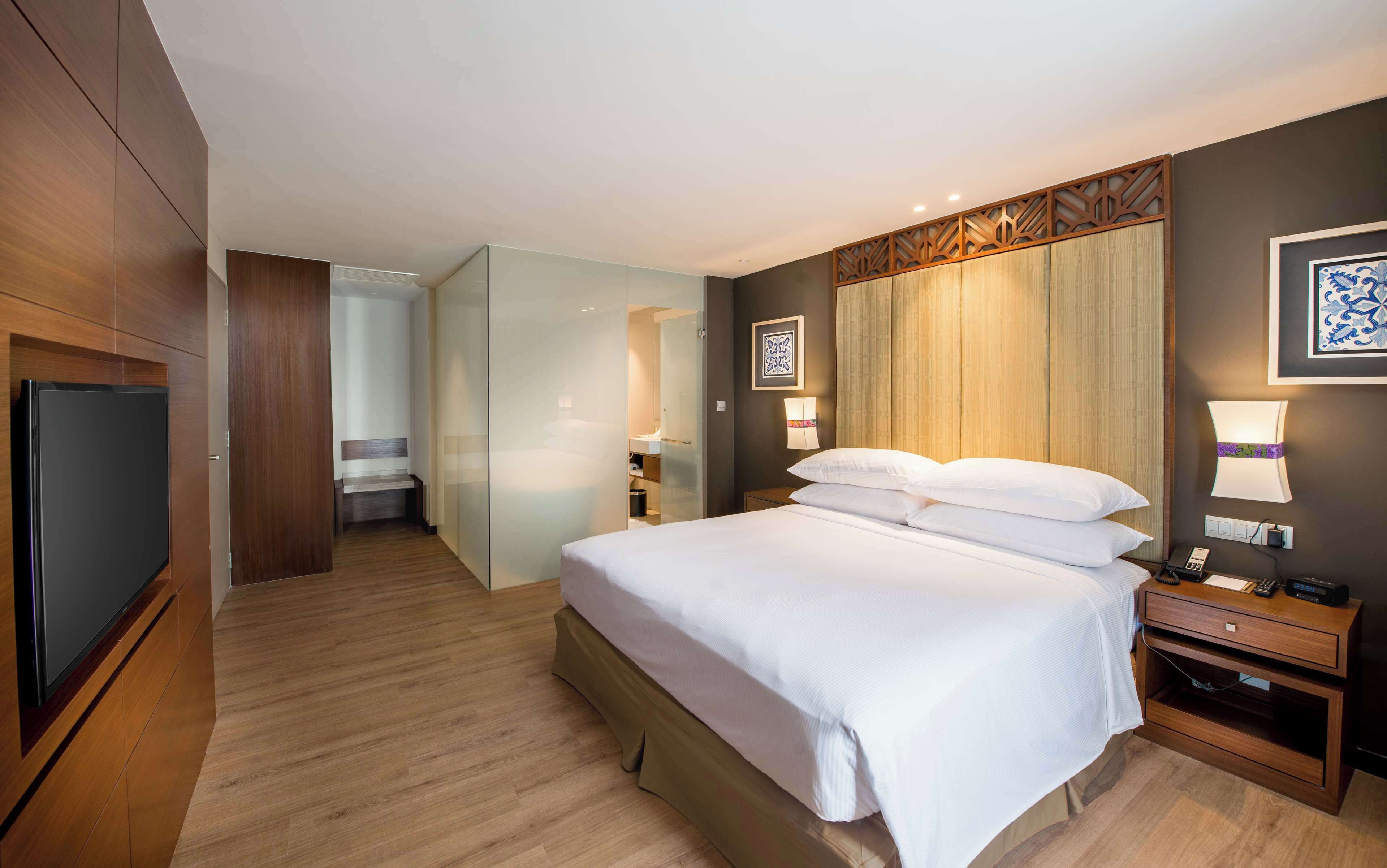A hotel room with a large bed, bedside tables, a TV, and a bathroom visible through a glass partition.