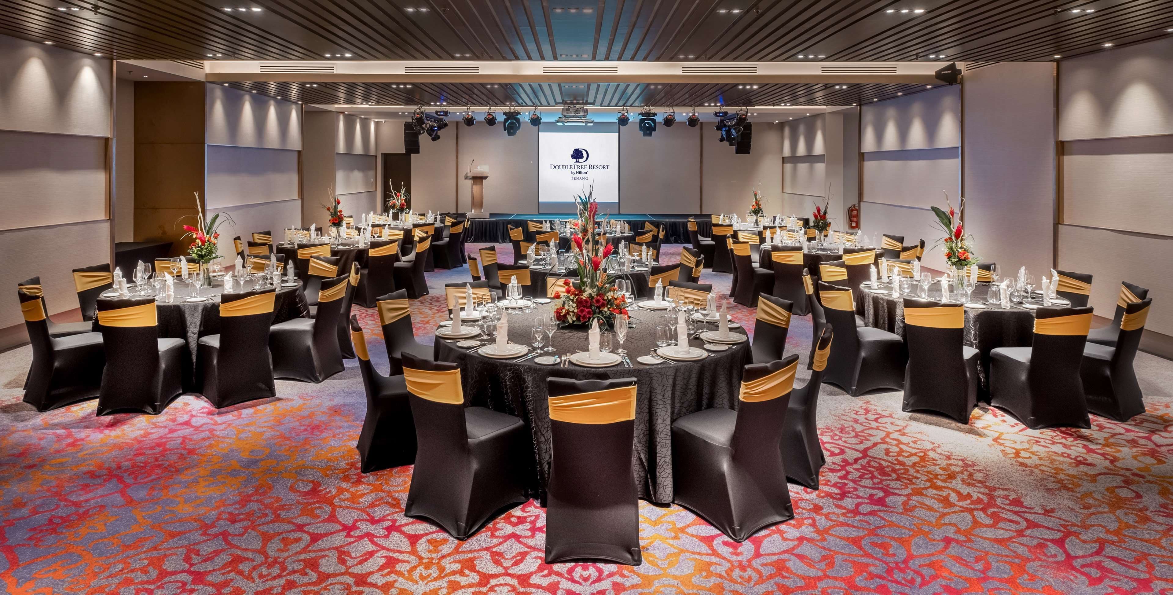 A conference room with multiple tables set for a formal event, featuring black chairs with gold sashes, a carpeted floor, and a projection screen in the background.