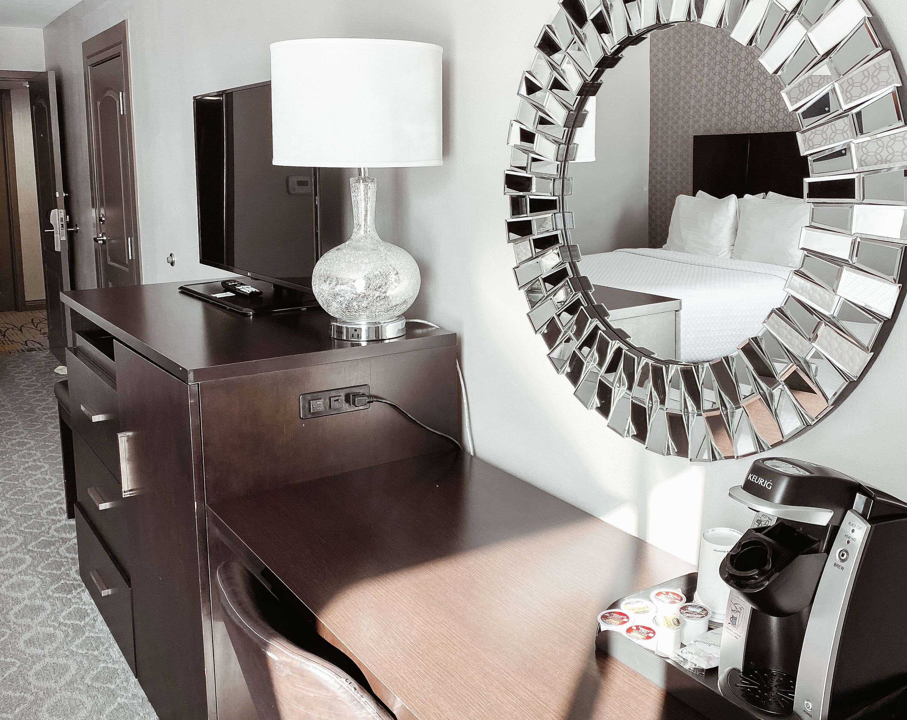 Habitación de hotel con minibar, espejo con diseño de mosaico y cama con ropa blanca.