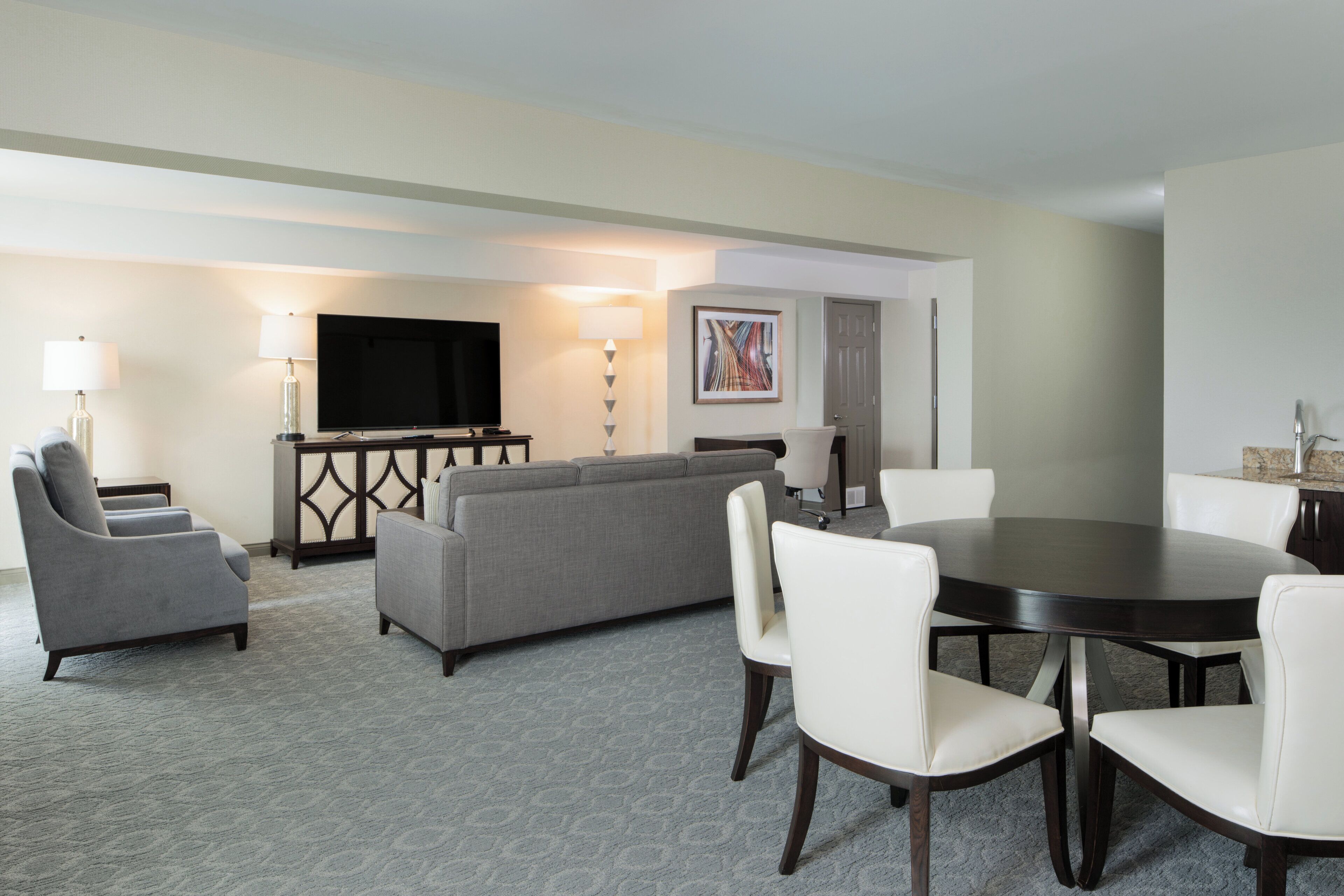 A hotel room with a gray sofa, two armchairs, a dining table with chairs, a flat-screen TV, a lamp, and a painting on the wall.