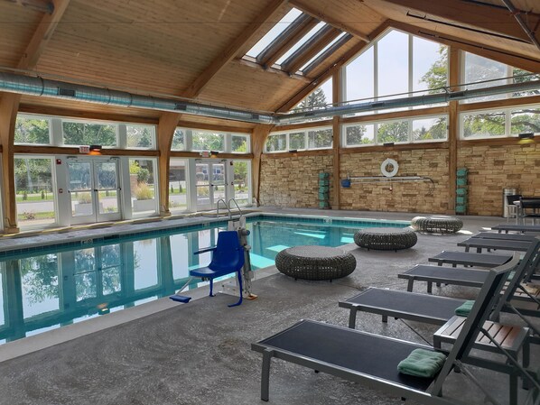 An indoor swimming pool with clear glass walls, a blue pool chair, and lounge chairs with green cushions.