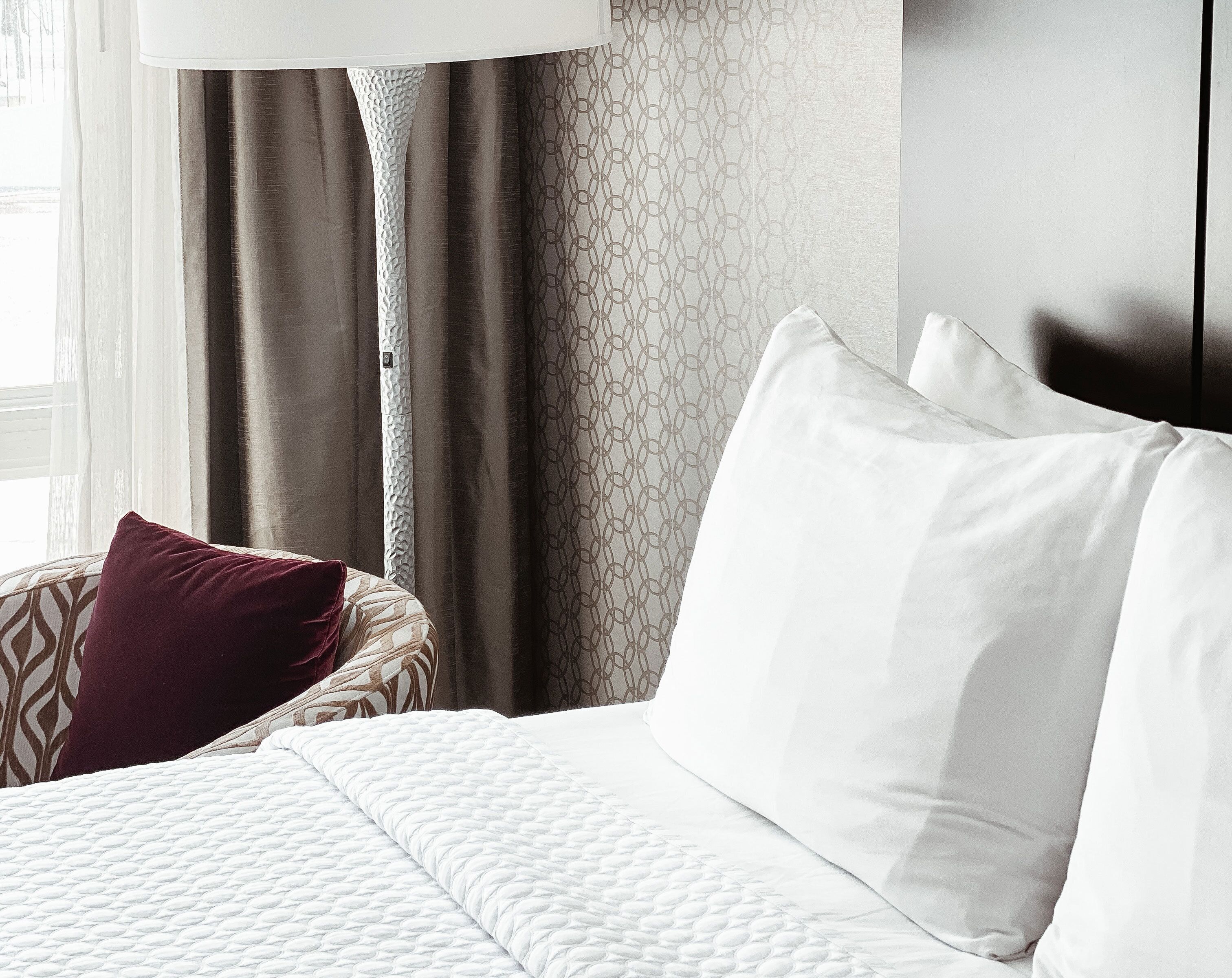 A hotel room with a bed, white bedding, a patterned pillow, and a floor lamp.