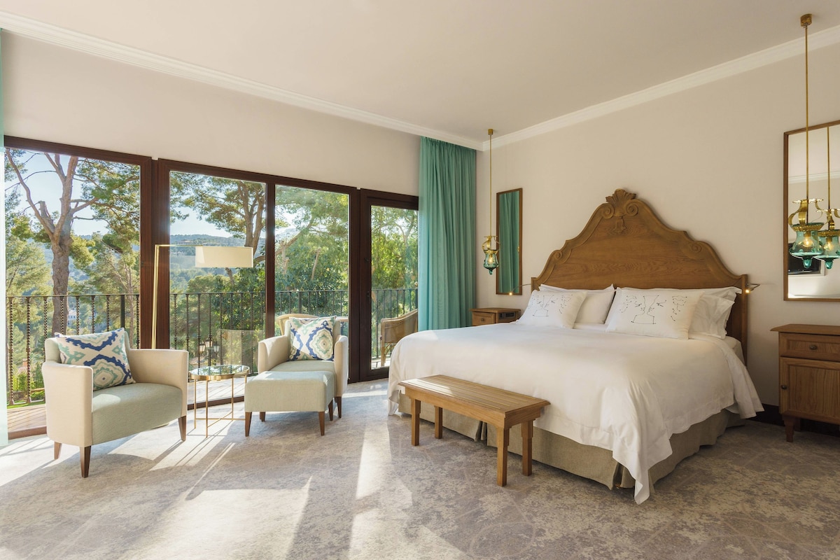 Une chambre spacieuse avec un grand lit, un banc, deux fauteuils et une vue sur l’extérieur grâce à de grandes fenêtres.