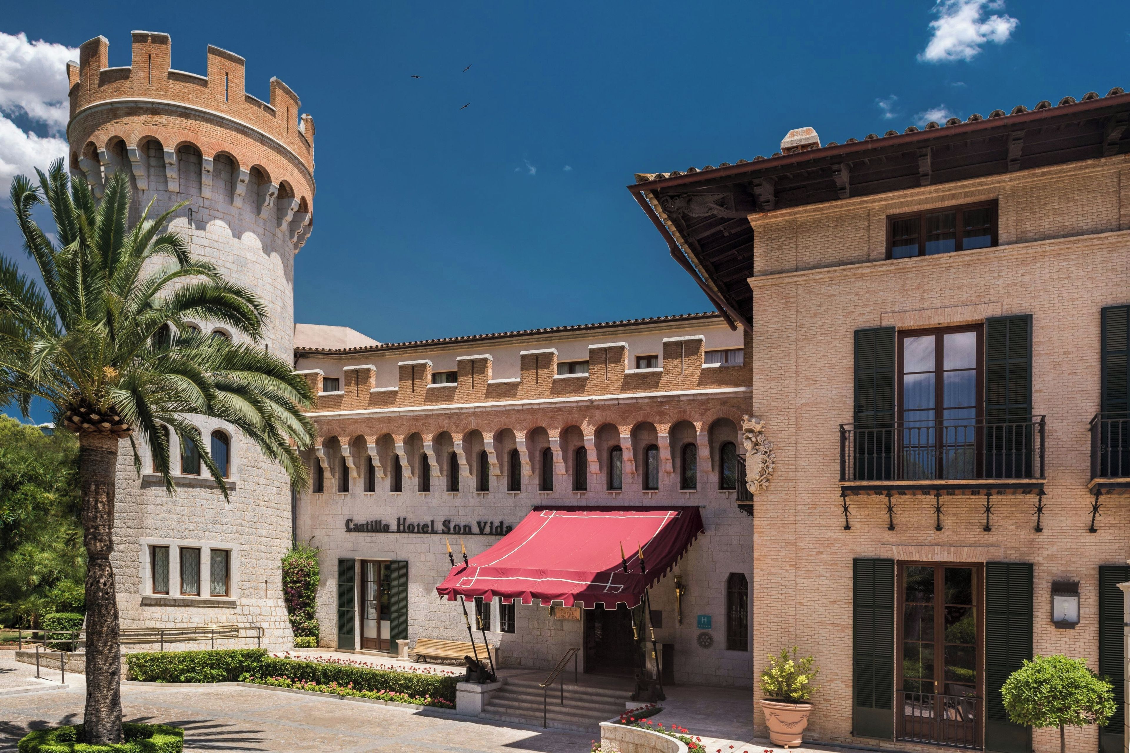 Un edificio histórico con una torre, un toldo rojo y una palmera.