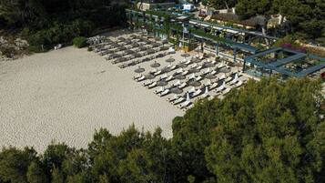 An aerial view of a beachside hotel with rows of sun loungers and umbrellas, surrounded by dense trees.