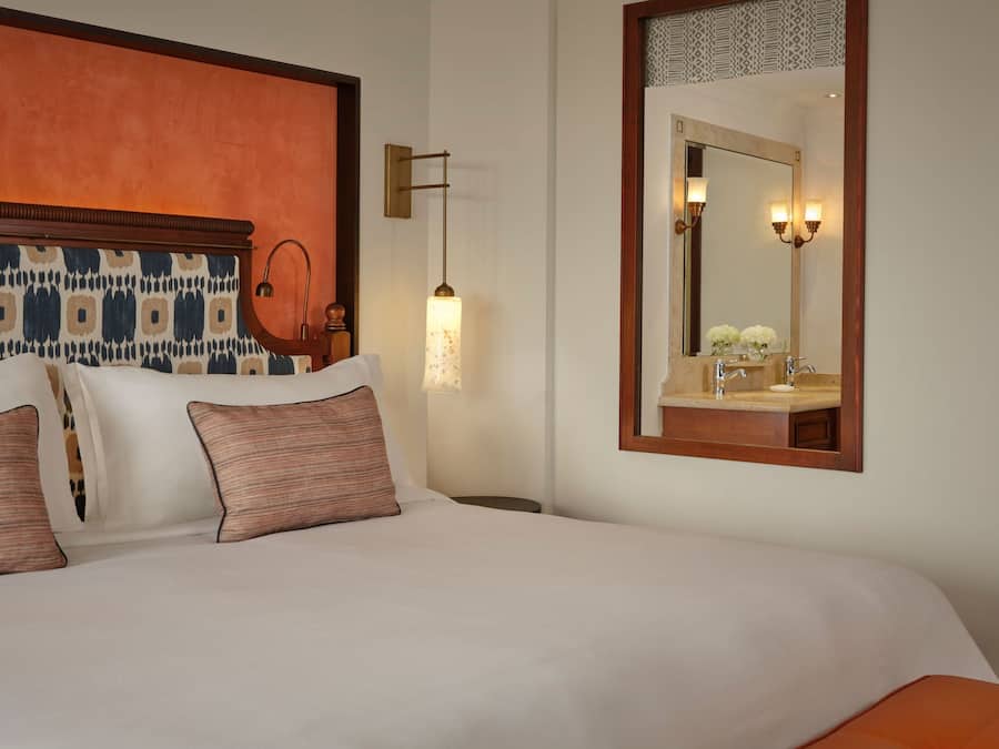 Habitación de hotel con cama, mesita de noche, lámpara, espejo y aplique de pared.