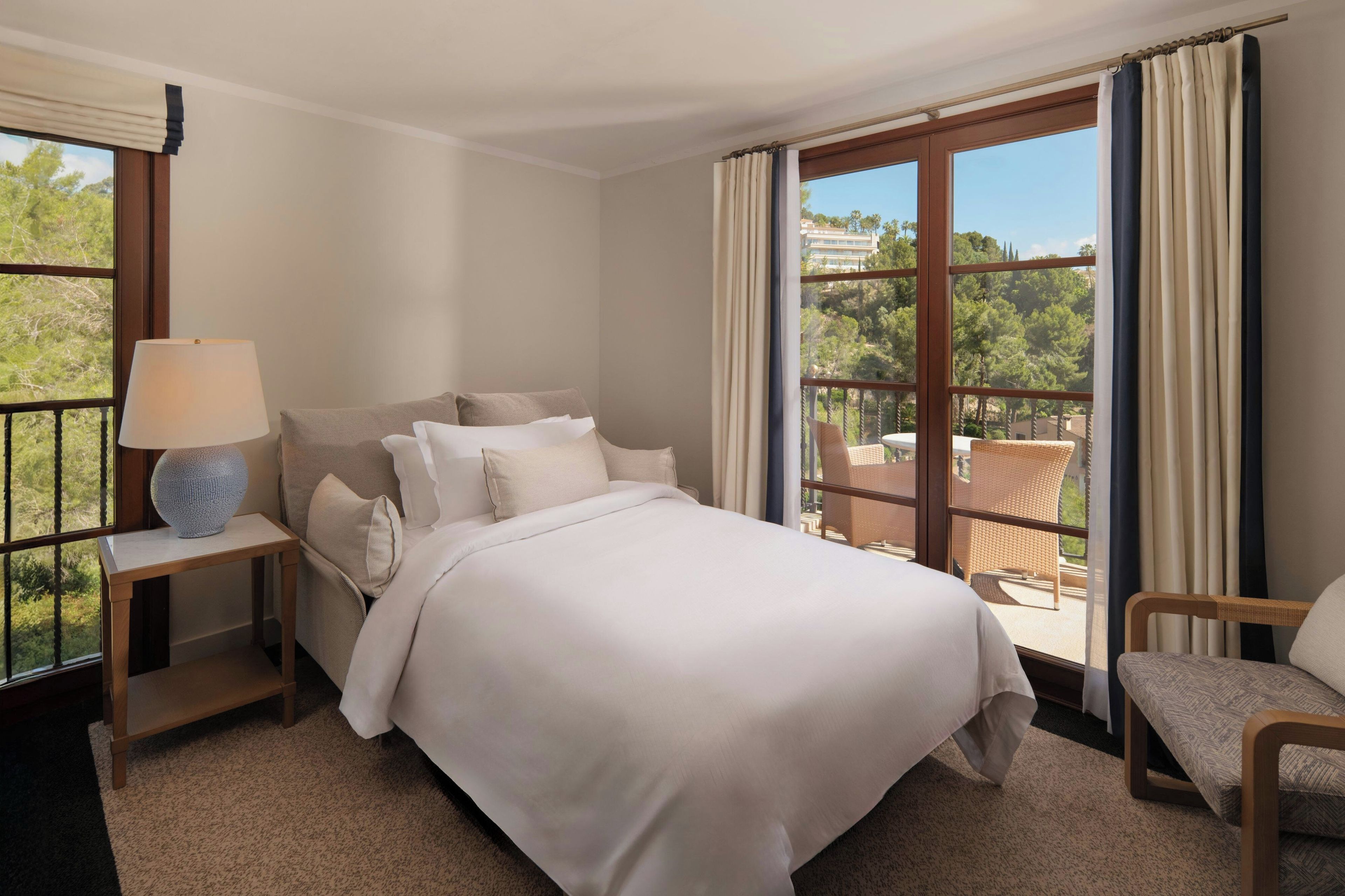 Habitación de hotel con cama, mesita de noche, lámpara, silla y vistas al exterior.
