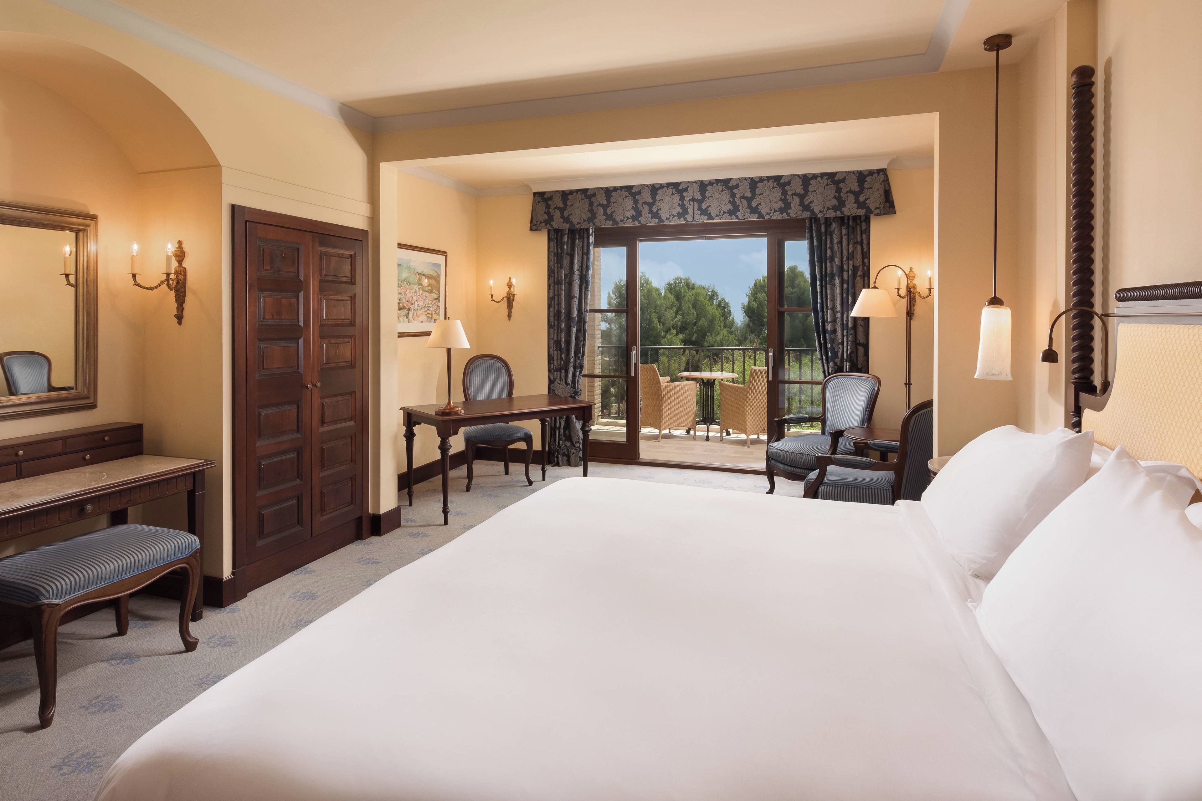 A hotel room with a large bed, a desk with a chair, a balcony with a table and chairs, and a view of the outdoors.
