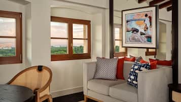A room with a view of a cityscape through large windows, a wicker chair, a sofa with patterned cushions, and a framed painting on the wall.