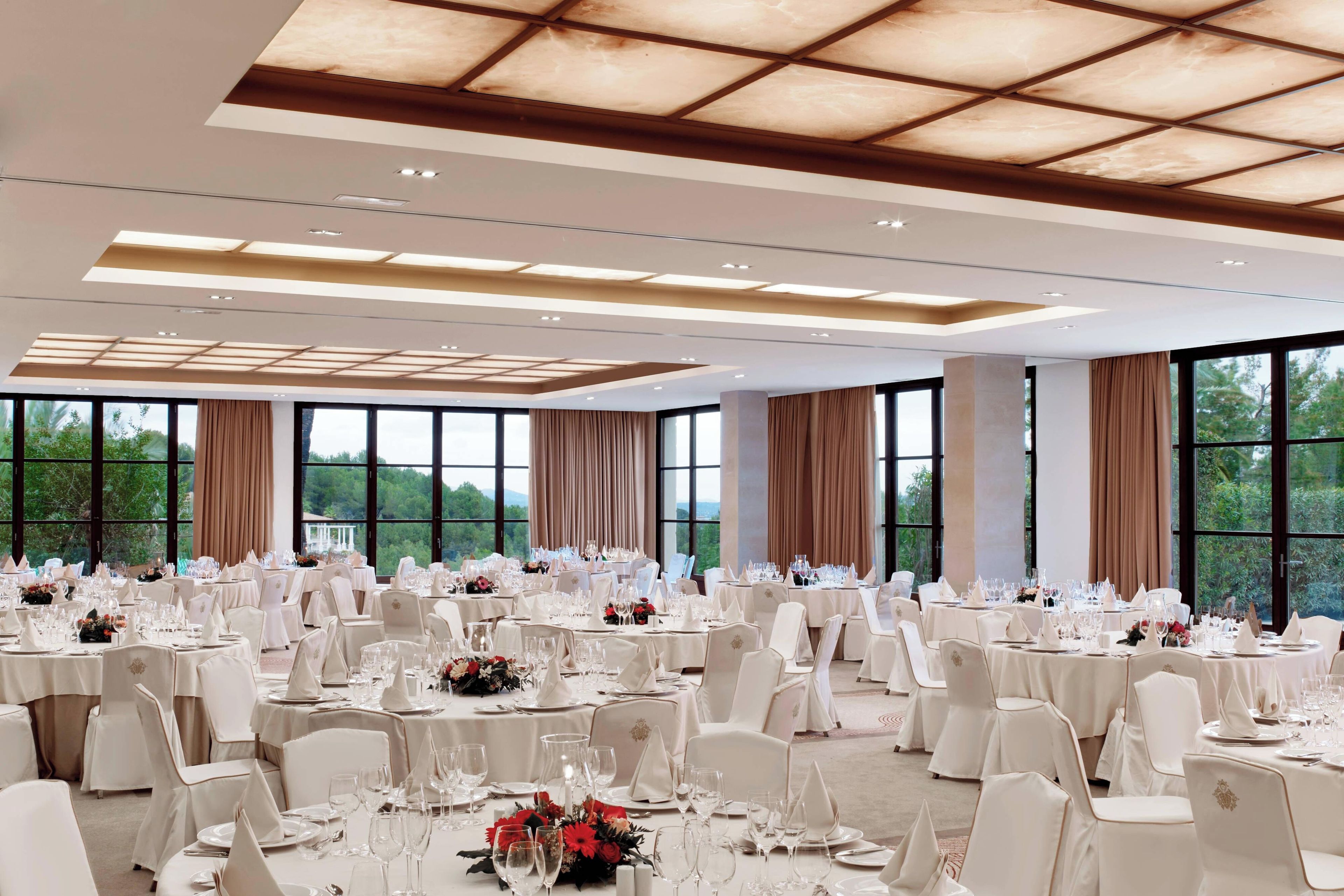 Um salão de banquetes amplo, com mesas redondas, cadeiras brancas e grandes janelas com vista para o exterior.