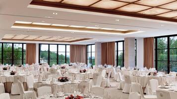 Um salão de banquetes amplo, com mesas redondas, cadeiras brancas e grandes janelas com vista para o exterior.