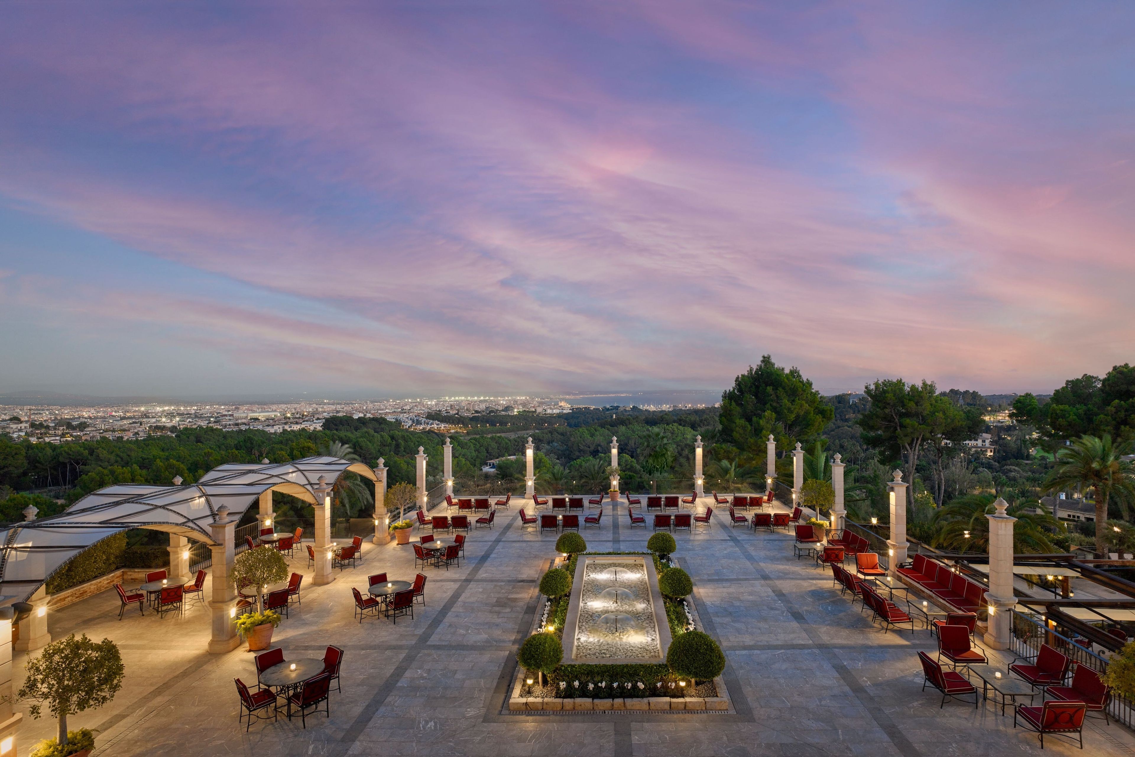 A rooftop terrace with red chairs and tables, a central water feature, and a view of the cityscape.