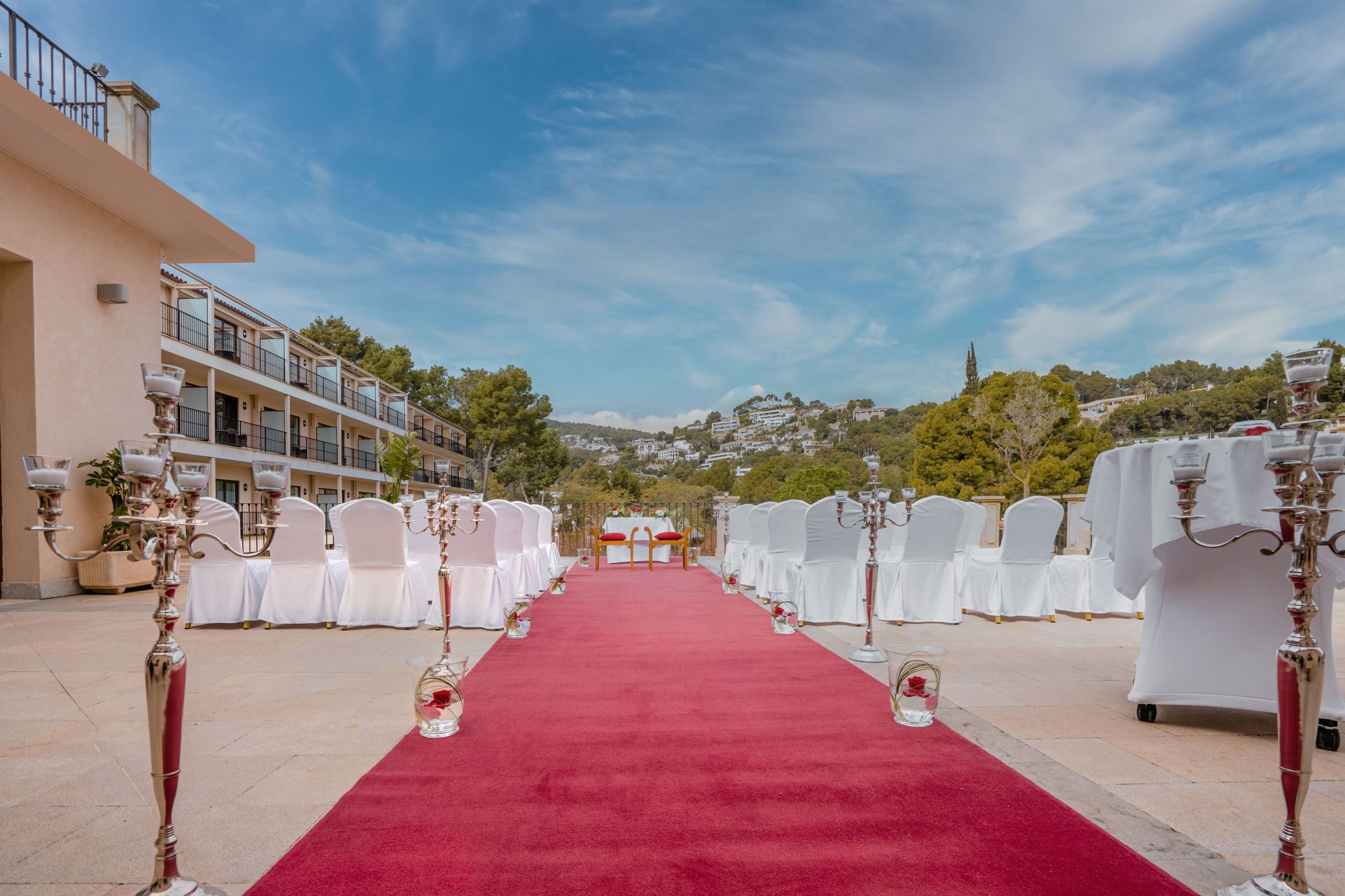 A red carpet aisle leading to a white altar with a backdrop of a building and trees.