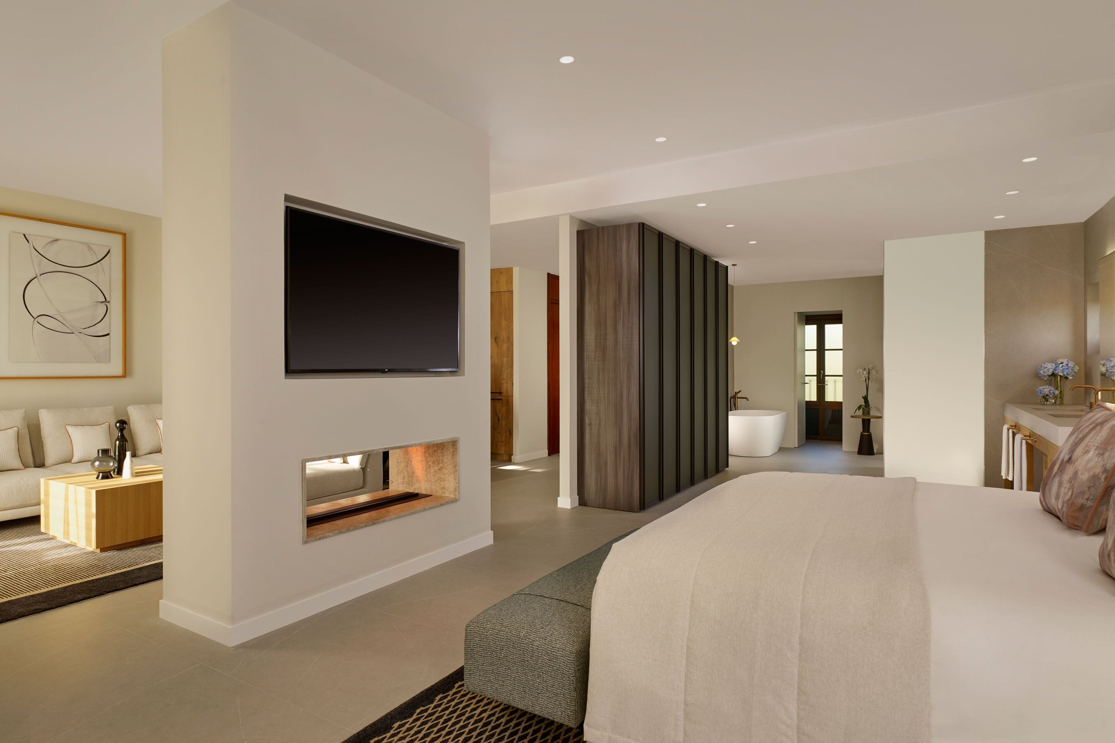 Habitació d'hotel moderna amb un llit gran, un televisor de pantalla plana penjat a la paret, una petita zona d'estar amb una taula de cafè, i un bany visible a través d'una porta oberta.