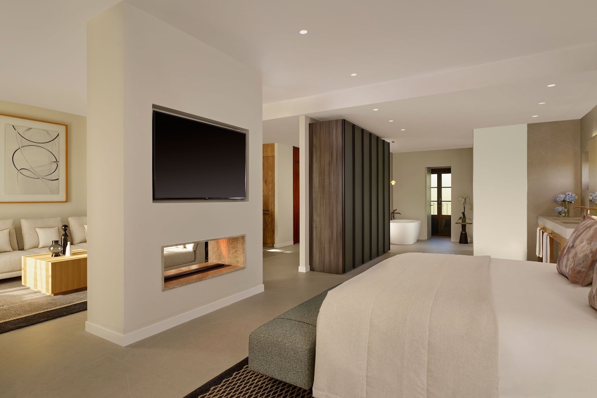 Une chambre d’hôtel moderne dotée d’un grand lit, d’un téléviseur à écran plat fixé au mur, d’un petit coin salon avec une table basse et d’une salle de bain visible à travers une porte ouverte.