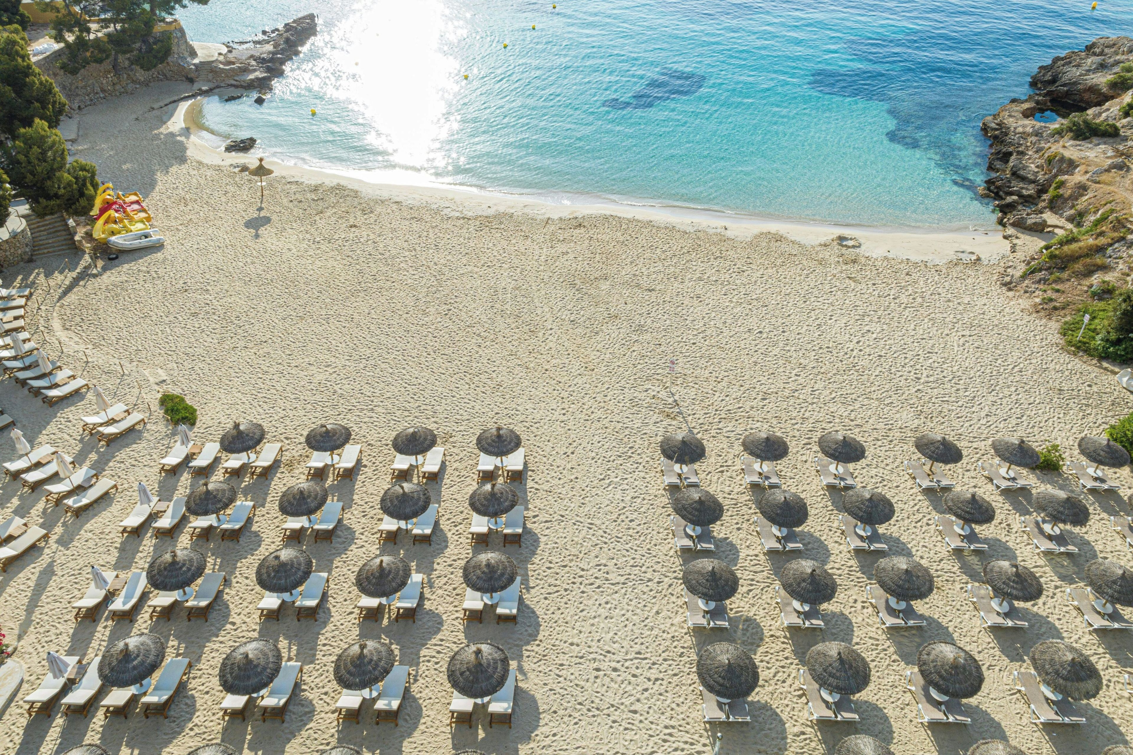 A beach with rows of sun loungers and umbrellas, a clear view of the sea, and rocky terrain.