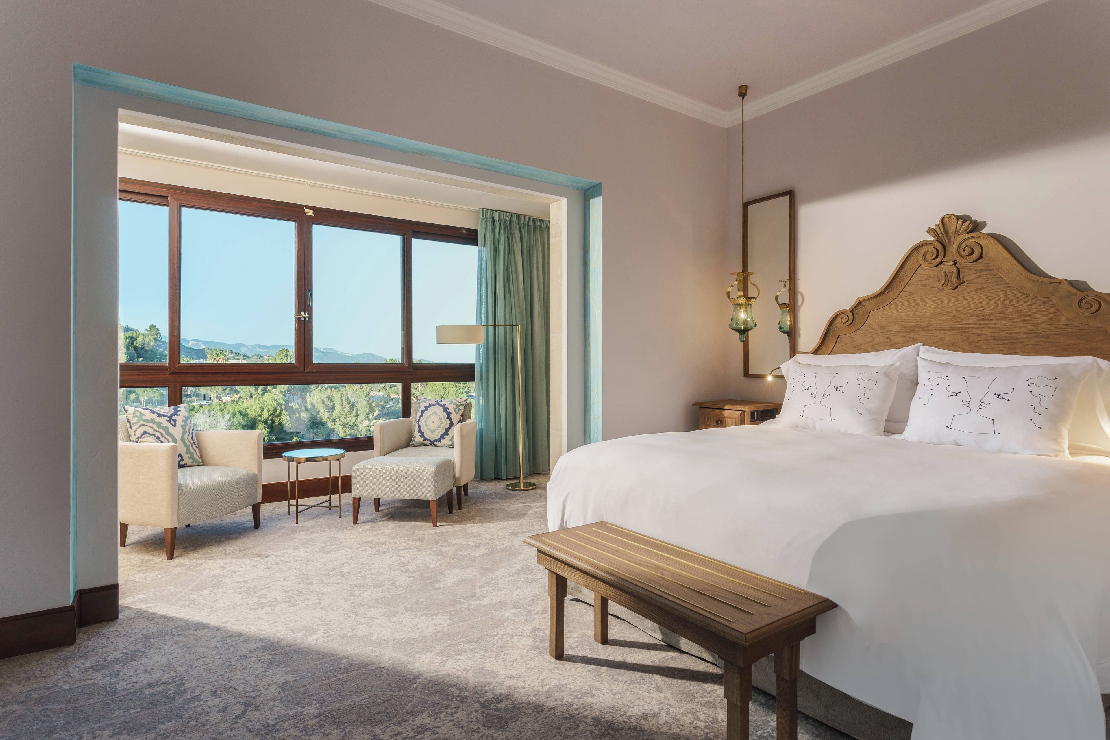 Une chambre d’hôtel avec un grand lit, une tête de lit en bois, un banc, deux fauteuils, une petite table et une vue sur l’extérieur grâce à une grande fenêtre.