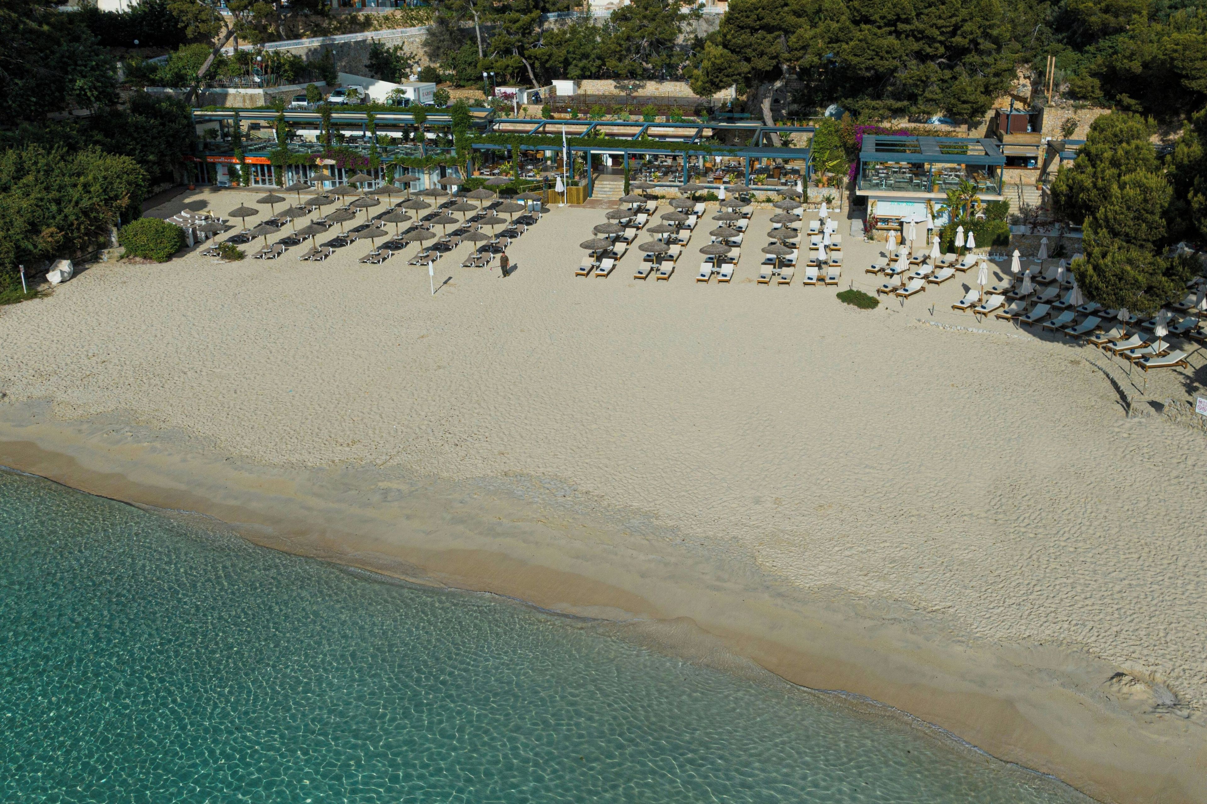 An aerial view of a beach with rows of sun loungers and umbrellas, a clear blue sea, and buildings in the background.