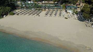 An aerial view of a beach with rows of sun loungers and umbrellas, a clear blue sea, and buildings in the background.
