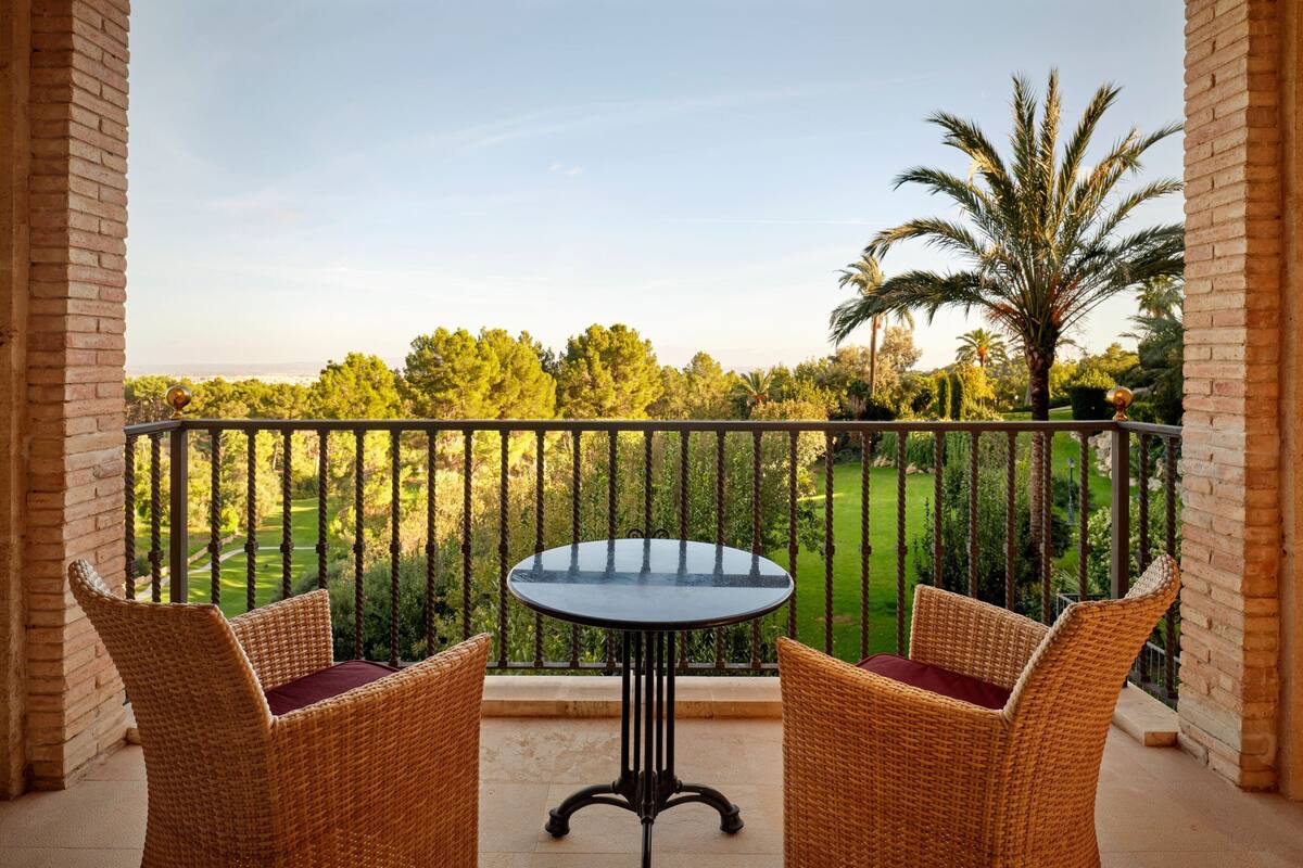Un balcon agrémenté de fauteuils et d’une table en osier, offrant une vue sur un jardin magnifiquement entretenu, avec des palmiers et un terrain de golf.