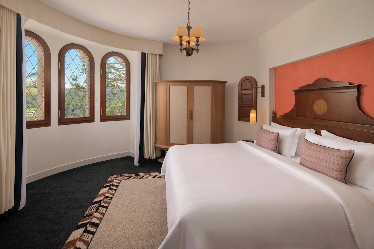Une chambre d’hôtel avec un grand lit, une armoire, un lustre et trois fenêtres en arc de cercle.