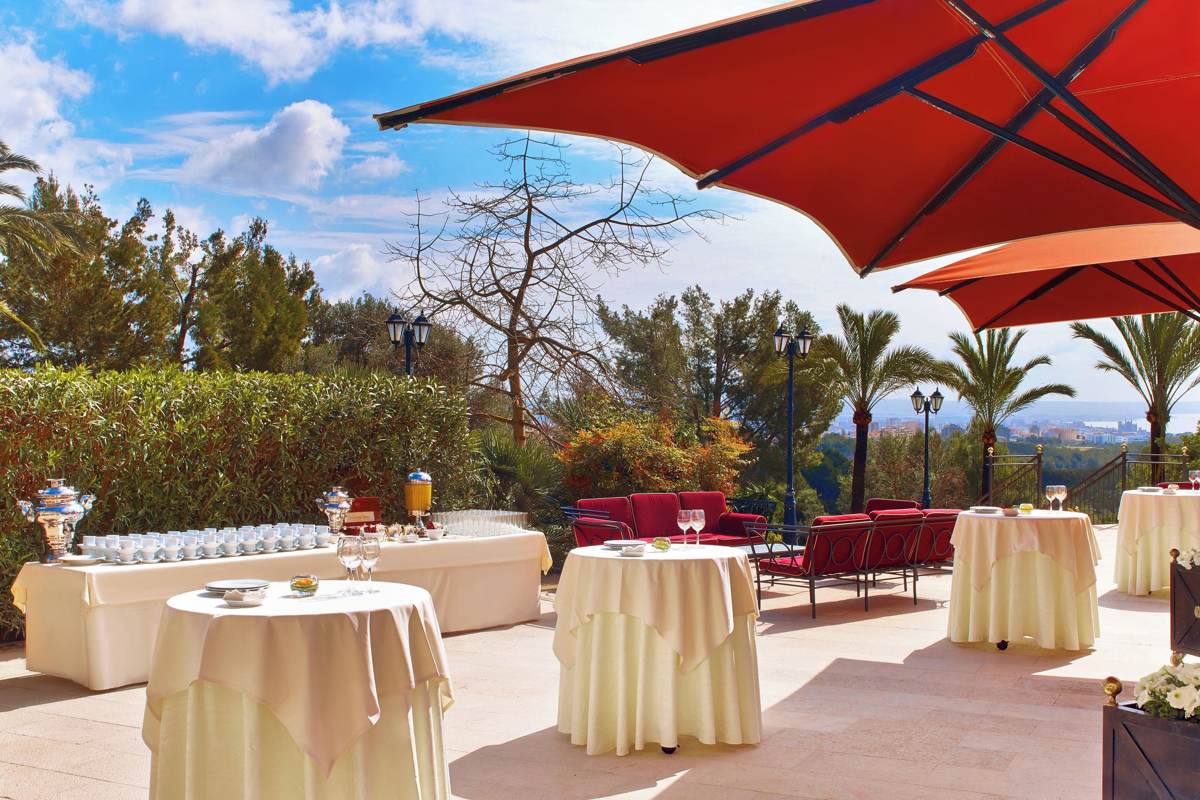 An outdoor dining area with round tables, red chairs, and a red umbrella providing shade.