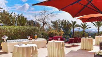 An outdoor dining area with round tables, red chairs, and a red umbrella providing shade.