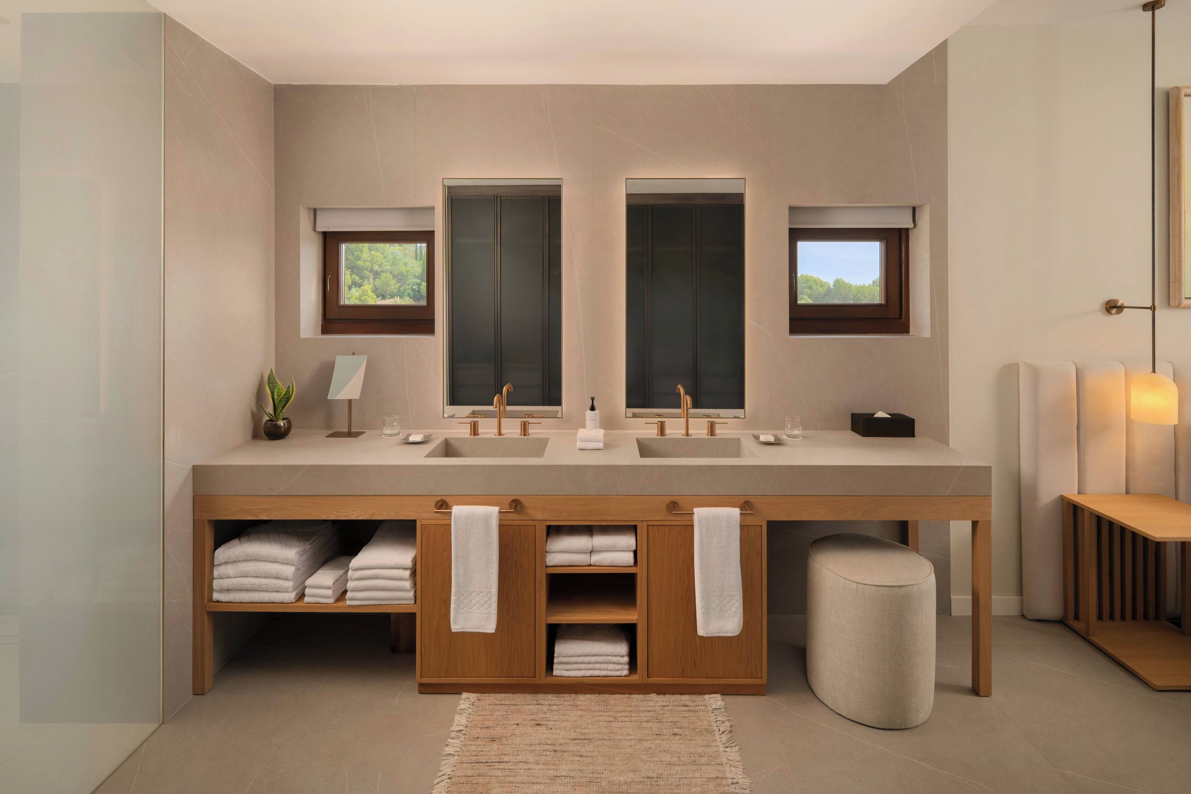 A modern bathroom with a large vanity, two sinks, and a mirror. There are towels and a plant on the vanity, and a small stool in front.
