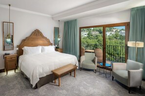Ein Hotelzimmer mit einem großen Bett, einem hölzernen Kopfteil, einer Bank, zwei Sesseln, einem kleinen Tisch und einem Balkon mit Blick auf die Stadt.