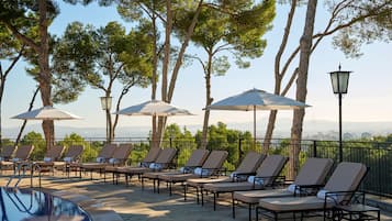 A poolside area with lounge chairs and umbrellas, surrounded by trees and a view of the mountains.