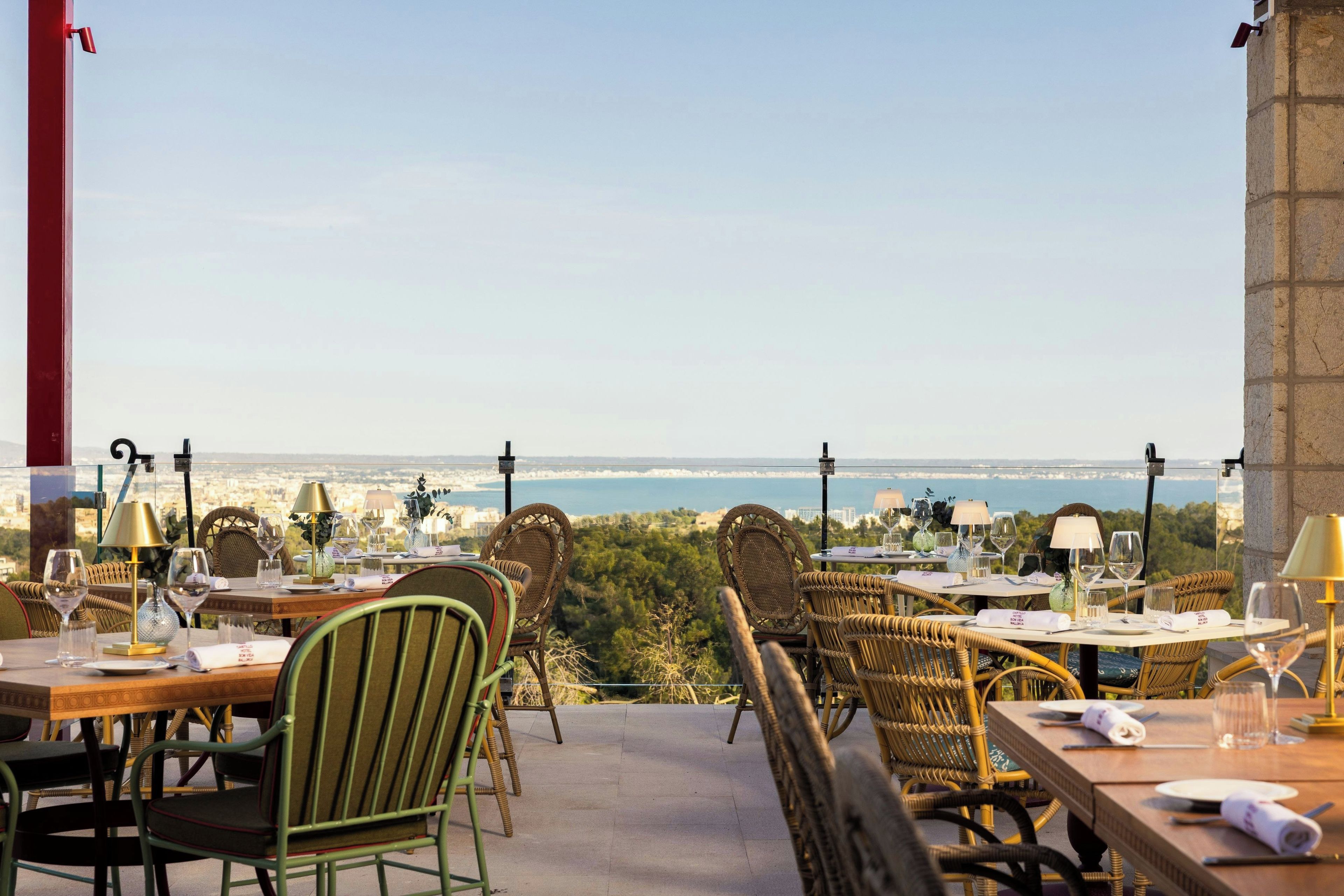 An outdoor dining area with wicker chairs and tables, overlooking a scenic view of the sea and cityscape.