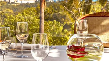 Mesa lista para comer con vasos transparentes, una jarrón de agua y platos, con vista a un paisaje urbano y palmeras.