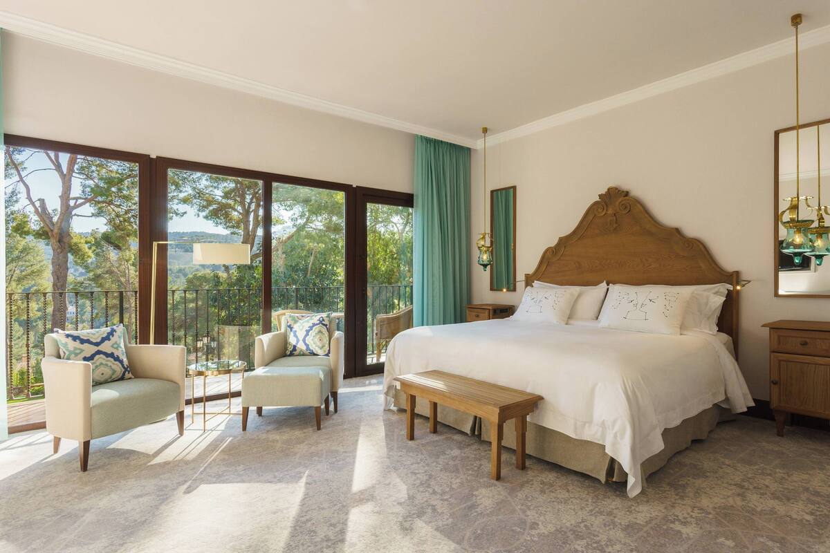 Une chambre spacieuse avec un grand lit, un banc, deux fauteuils et une vue sur l’extérieur grâce à de grandes fenêtres.