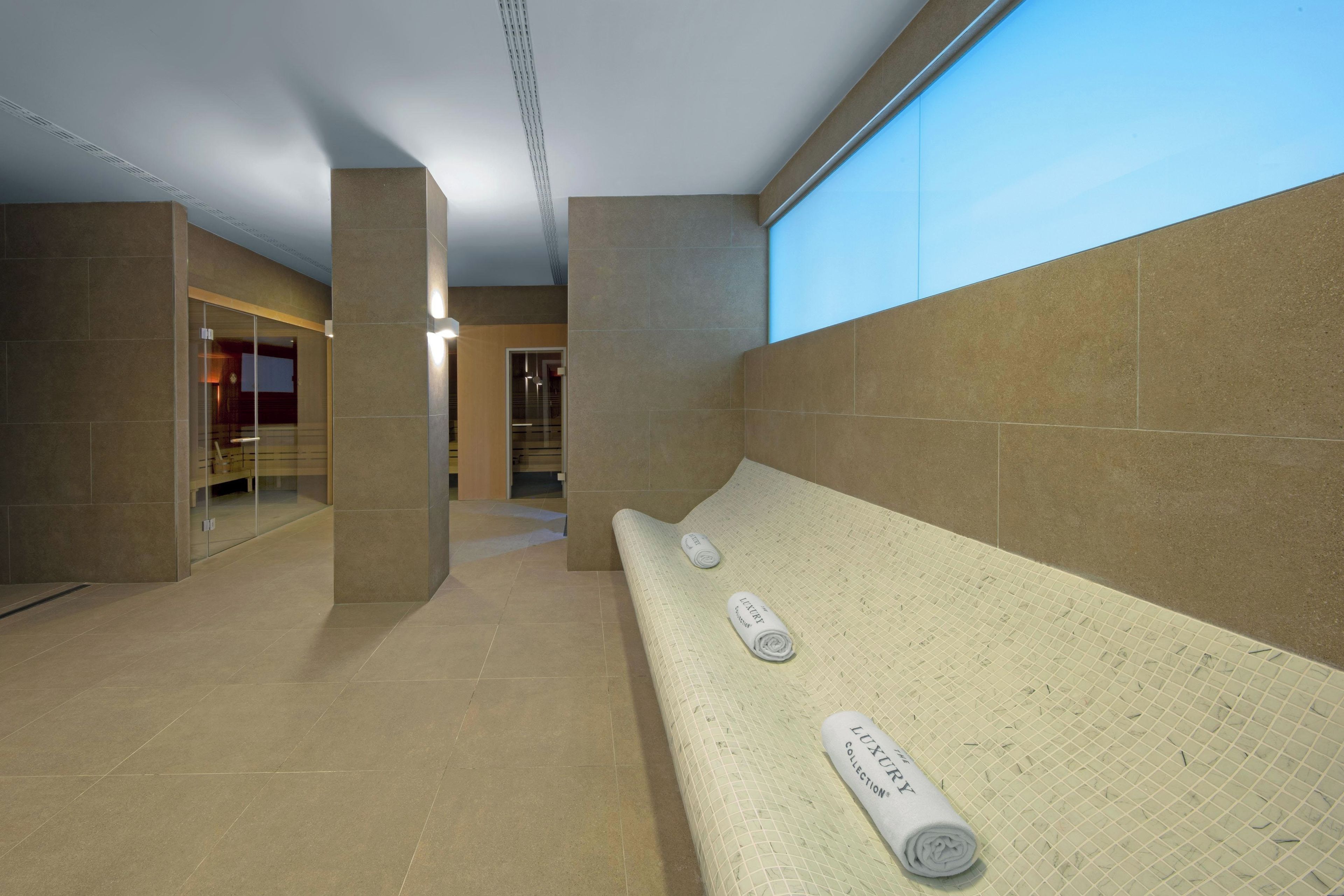 A modern interior with a long bench, two rolled towels, and a glass door leading to another room.