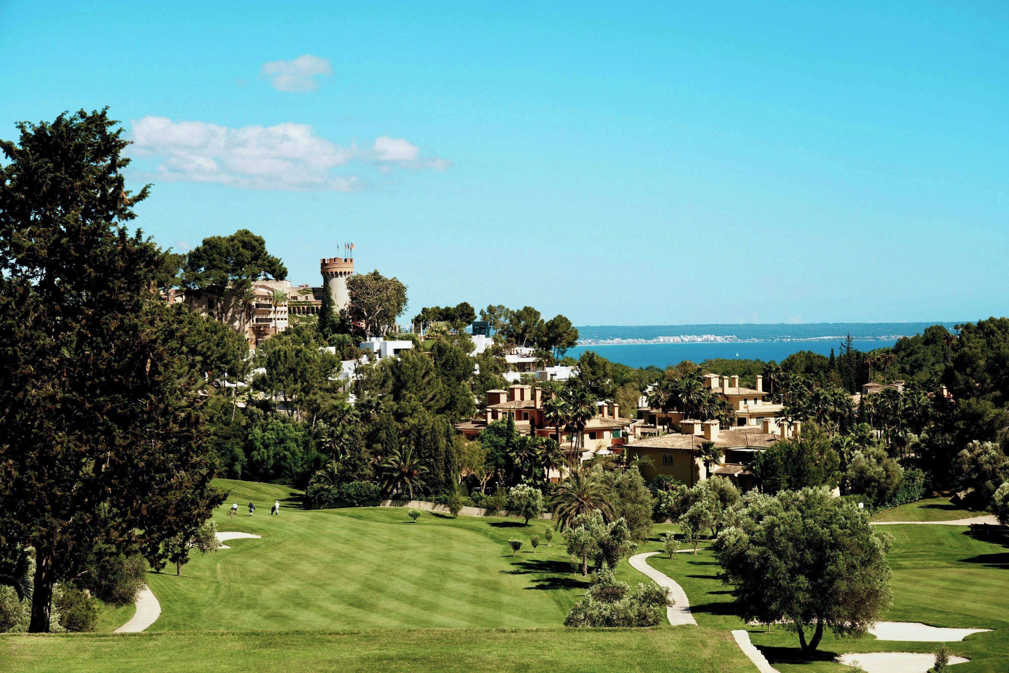 A golf course with a castle-like structure in the background, surrounded by lush greenery and a clear view of the sea.