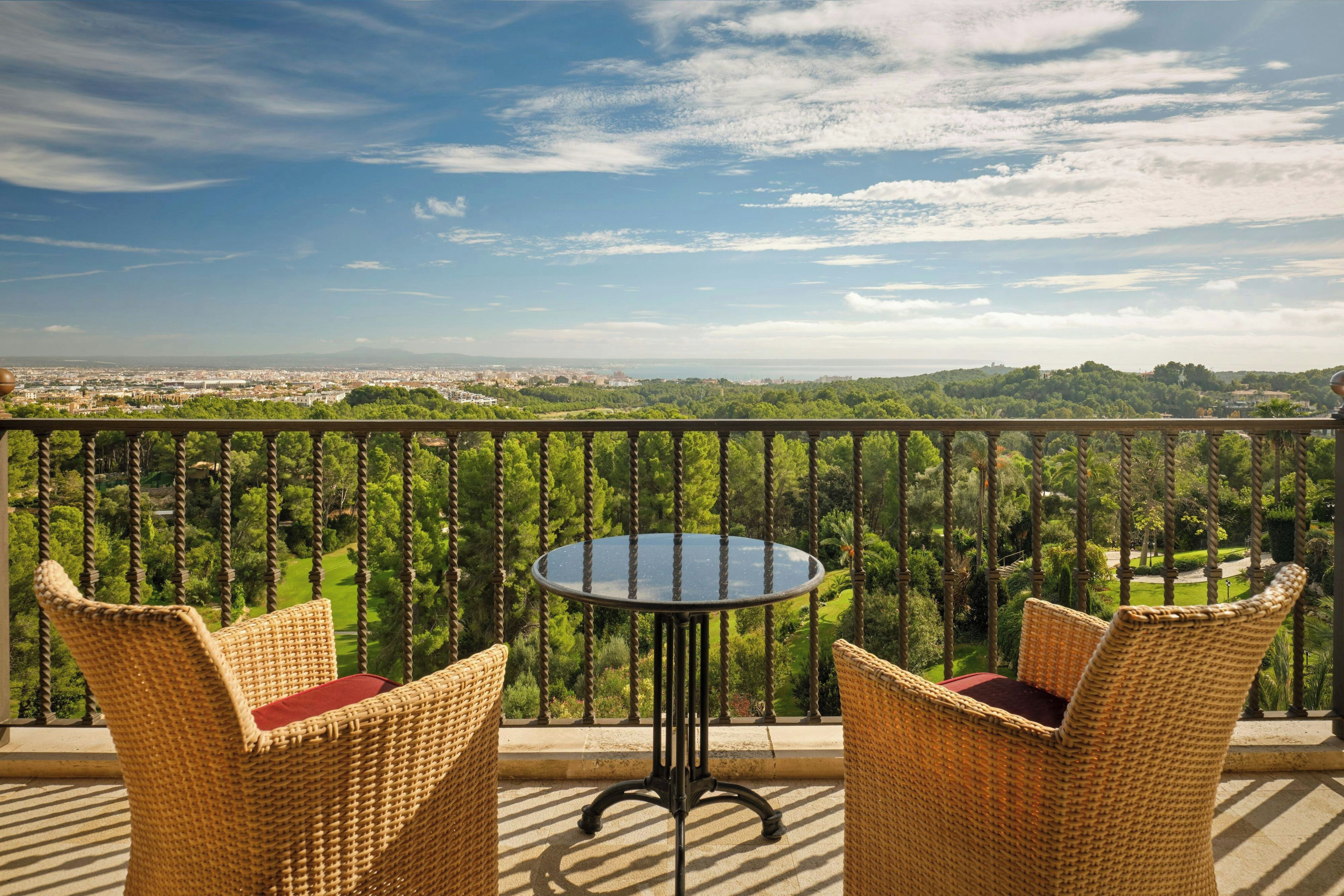 Un balcón con sillas de mimbre y una mesa de vidrio, con vistas a un paisaje urbano y vegetación.