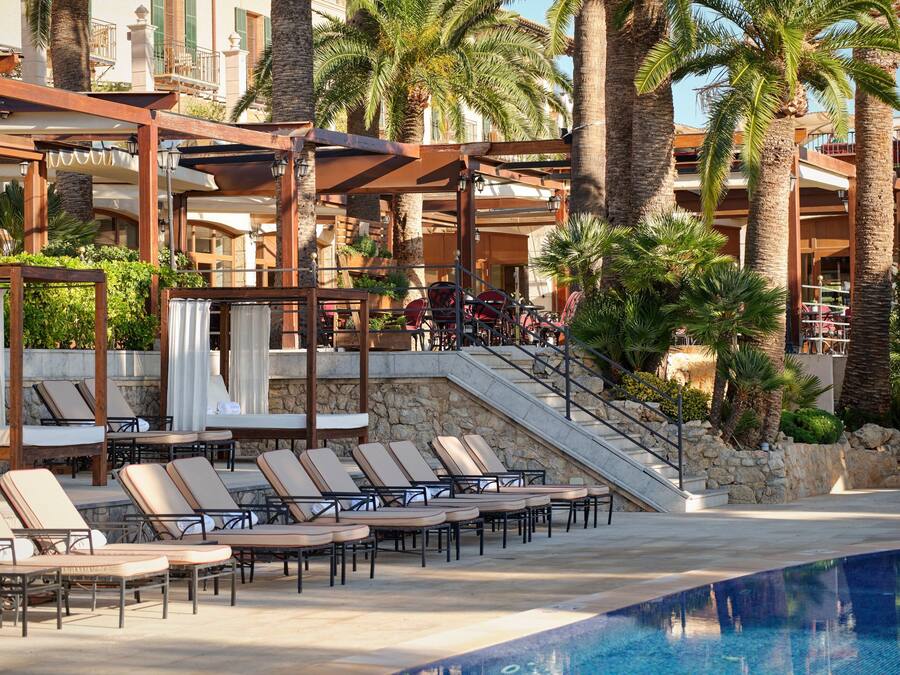 Zona de piscina con sillones de descanso y una alberca, rodeada de palmeras y un edificio con balcones.