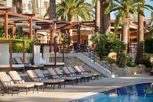 Zona de piscina con sillones de descanso y una alberca, rodeada de palmeras y un edificio con balcones.
