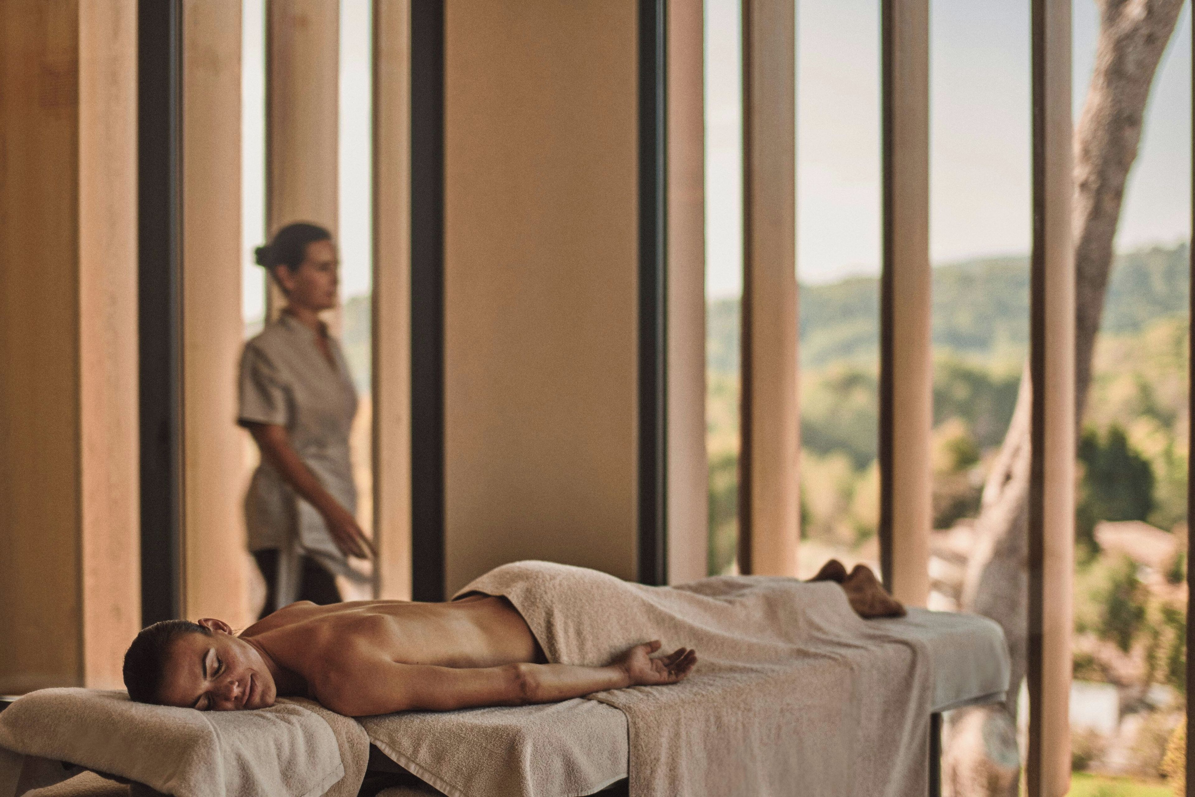 Une salle de massage où une personne est allongée sur une table de massage, une autre personne se tient à l’arrière-plan, et on peut admirer un paysage naturel à travers de grandes fenêtres.